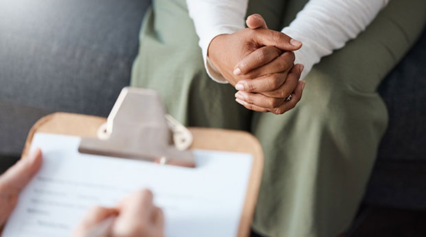 A closeup of a person's hands folded. In front of their hands is someone writing on a clipboard.